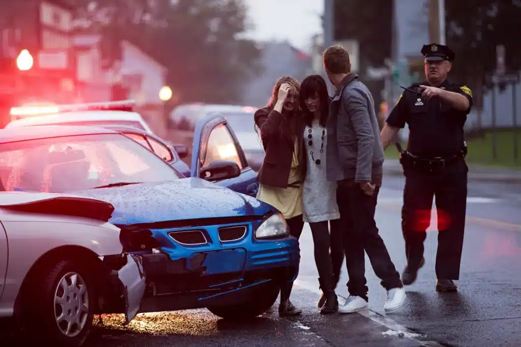 Two women and a man stand near a blue car with front-end damage, while a police officer gestures nearby. The scene is outdoors on a rainy day.