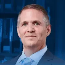 A professional headshot of Christopher DiBella against a blue background, wearing a suit and tie.