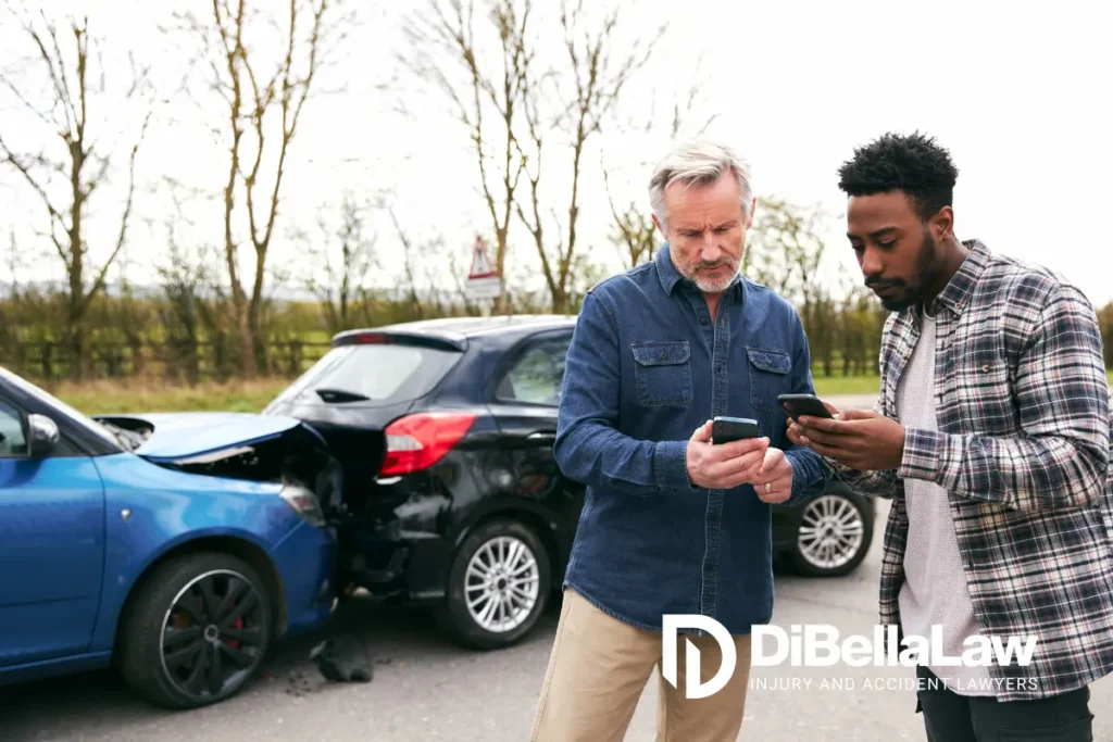 Two men standing outside on a road looking down at their phones, with a rear-end car accident visible behind them.